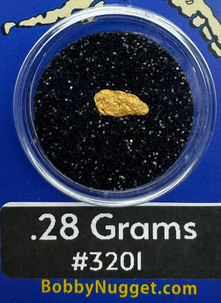 Gold nugget in a clear container with black sand on a blue background, labeled '.28 Grams #3201' and 'BobbyNugget.com'.