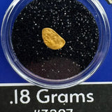 Gold nugget in a clear container with black sand, labeled '.18 Grams #3207' on a blue background.