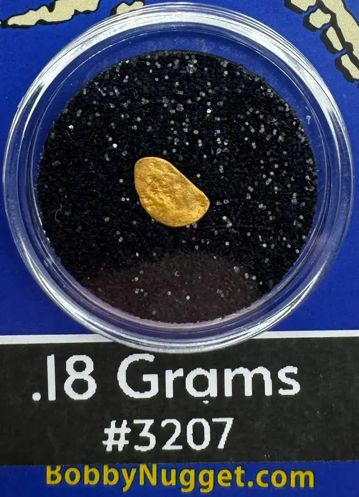 Gold nugget in a clear container with black sand, labeled '.18 Grams #3207' on a blue background.